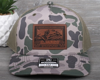 May include: A camouflage trucker hat with a brown leather patch featuring a vintage duck hunting stamp. The patch reads "U.S. Department of Agriculture Migratory Bird Hunting Stamp". The hat is adjustable and has a mesh back.