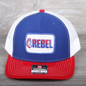 May include: A blue and white trucker hat with a red bill. The hat has a white mesh back and a red, white, and blue patch that says "Rebel" on it. The hat is adjustable and has a Richardson logo on the side.