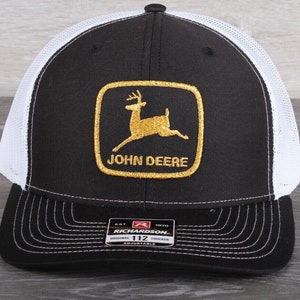 May include: Black and white trucker hat with a gold John Deere logo featuring a leaping deer. The logo is on a green square with a gold border. The hat is adjustable and has a white mesh back.