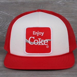May include: A red and white trucker hat with a red square patch that says "Enjoy Coke" in white lettering. The hat has a mesh back.