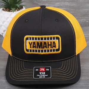 May include: Black and yellow Yamaha trucker hat. The front panel is black with a yellow embroidered Yamaha logo. The mesh back is yellow. The hat has a curved brim and an adjustable snap closure.