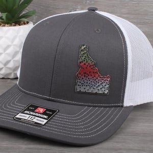 May include: A gray trucker hat with a white mesh back and a curved brim. The hat features a colorful patch of the state of Idaho with a fish design. The hat is adjustable and has a Richardson 112 tag.