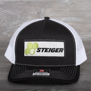 May include: Black and white trucker hat with a white mesh back. The front of the hat features a black patch with a green logo that says "Steiger" and a green "S". The hat also has a black label that says "Richardson 112 Original Trucker Adjustable".