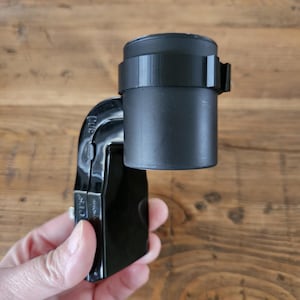 Puede incluir: Soporte de cámara negro con un accesorio cilíndrico negro. El soporte tiene un pequeño botón plateado y el texto "CIS" impreso en él.