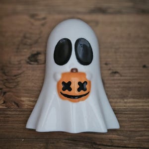 May include: White ghost figurine holding a small orange pumpkin with a black smiling face and two black X's for eyes.