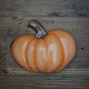 May include: A small, orange ceramic pumpkin with a brown stem. The pumpkin is sitting on a wooden surface.