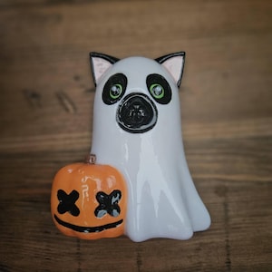 May include: A white ghost figurine with black cat ears and green eyes holds an orange pumpkin with a black 'XX' face.