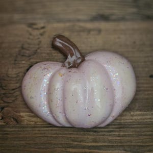 Puede incluir: Una pequeña calabaza de cerámica rosa con un tallo marrón. La calabaza está decorada con purpurina iridiscente.