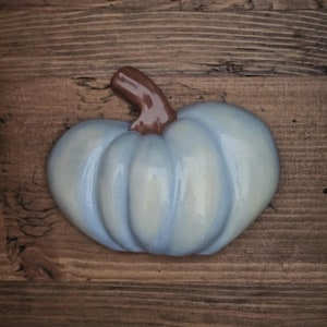 May include: A light blue ceramic pumpkin with a brown stem. The pumpkin is sitting on a wooden surface.