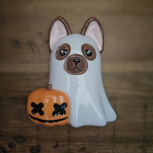 May include: A white ghost dog figurine with brown ears and a brown nose, holding a small orange pumpkin with black X's for eyes. The figurine is on a wooden surface.