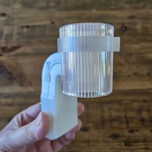 May include: A white plastic night light with a clear plastic shade. The shade has a textured surface and a white band around the middle.