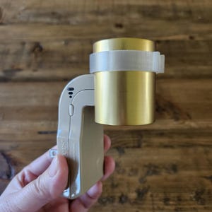 May include: A beige and white electric outlet adapter with a gold-coloured cylindrical device attached to it. The device is secured with a white plastic strap.