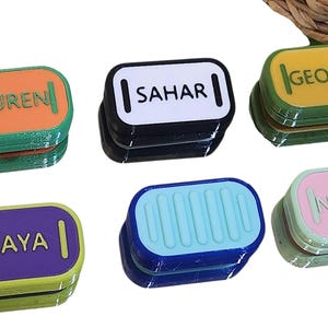 以下が含まれることがあります： さまざまな色のパーソナライズされたネームタグのコレクション。各タグには、Lauren、Sahar、George、Maya、Normaなど、異なる名前が記載されています。タグは角が丸い長方形で、積み重ねられています。