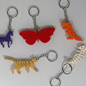 Puede incluir: Seis llaveros diferentes con una variedad de diseños. Los llaveros incluyen un unicornio morado, una mariposa roja, un dinosaurio naranja, un esqueleto de pez blanco y un gato amarillo.