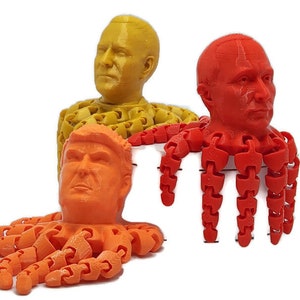 Puede incluir: Tres figuras articuladas impresas en 3D con rostros humanos. Una es naranja, otra roja y otra amarilla. Cada figura tiene una cabeza y múltiples apéndices en forma de tentáculos. Las figuras están sobre un fondo blanco.
