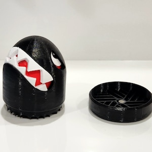 Puede incluir: Una figura impresa en 3D en blanco y negro de un personaje del videojuego Super Mario Bros. El personaje es negro, redondo y tiene una sonrisa blanca y dentada. La figura est&aacute; separada en dos partes, una cabeza y una base.