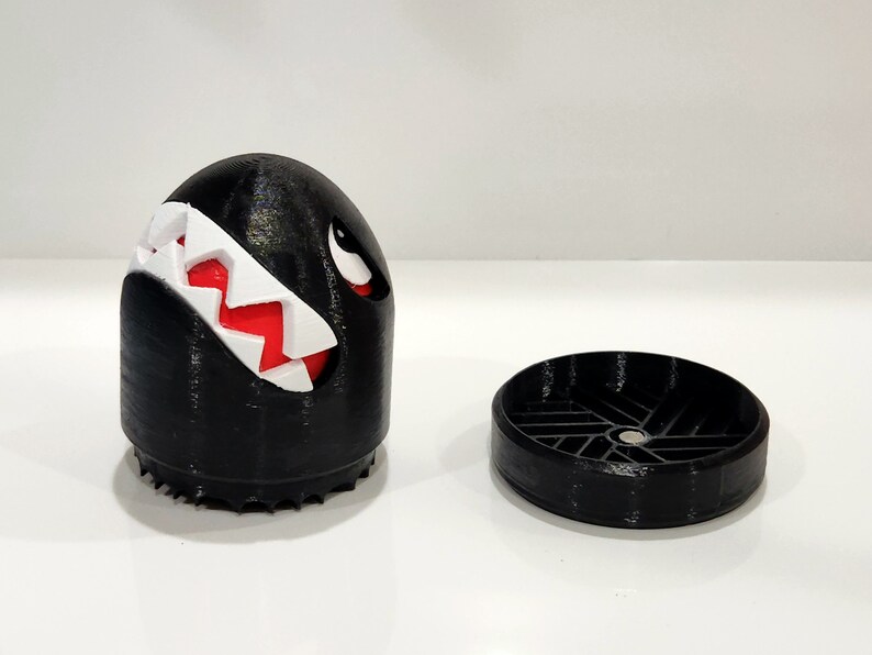 Puede incluir: Una figura impresa en 3D en blanco y negro de un personaje del videojuego Super Mario Bros. El personaje es negro, redondo y tiene una sonrisa blanca y dentada. La figura est&aacute; separada en dos partes, una cabeza y una base.