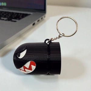May include: A black Bullet Bill keychain with a silver keyring. The keychain features a cartoon-style design with white eyes and a red and white mouth. The keychain is made of a 3D-printed material.
