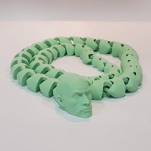 Può includere: Una collana verde stampata in 3D con un ciondolo a forma di testa umana. La collana è composta da pezzi che si incastrano per formare una catena.