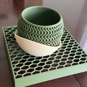 May include: A sage green cylindrical planter with a textured wavy pattern on the exterior, nested inside a smooth cream-colored bowl, all resting on a flat, square, sage green base featuring a repeating hexagonal grid cutout pattern.