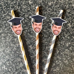 May include: Three graduation-themed paper straws. Each straw features a smiling face wearing a mortarboard. The straws are striped in gold, copper, and grey, with white stripes. The background is a speckled grey surface.