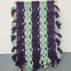 May include: A macrame wall hanging with a geometric pattern in shades of purple and green. The hanging has tassels and four small bells at the bottom.