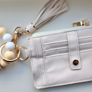 May include: A cream-colored wristlet keychain with a beaded bracelet, a matching tassel, and a small cardholder wallet. The bracelet features white and beige beads. The wallet has multiple card slots and a gold-tone zipper.