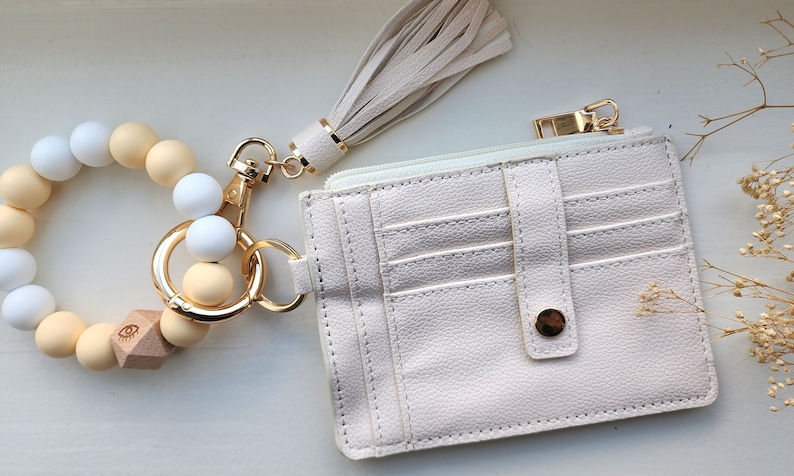 May include: A cream-colored wristlet keychain with a beaded bracelet, a matching tassel, and a small cardholder wallet. The bracelet features white and beige beads. The wallet has multiple card slots and a gold-tone zipper.