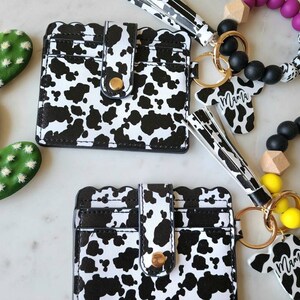 May include: A collection of accessories featuring a cow print design. Includes two cardholders, a keychain with a cow-shaped charm that says "Mama", and two beaded bracelets. The cardholders have a scalloped edge and a gold snap closure.