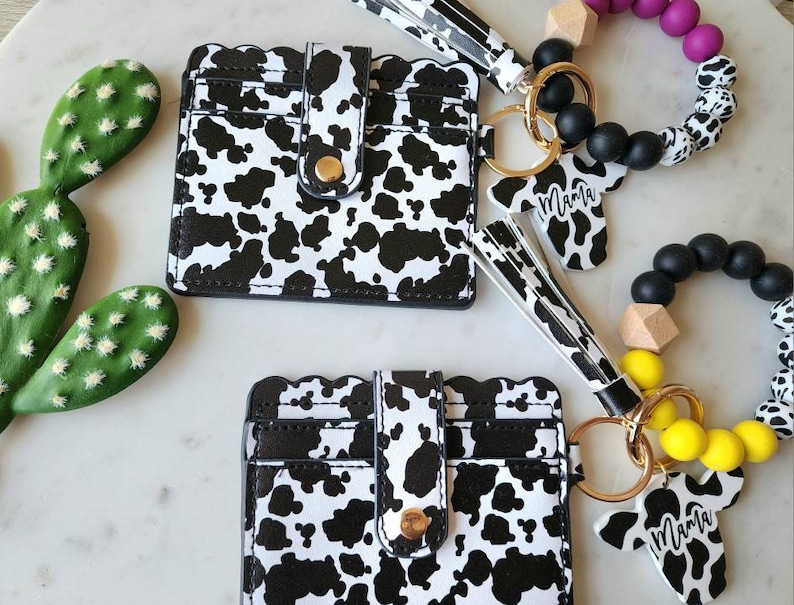 May include: A collection of accessories featuring a cow print design. Includes two cardholders, a keychain with a cow-shaped charm that says "Mama", and two beaded bracelets. The cardholders have a scalloped edge and a gold snap closure.