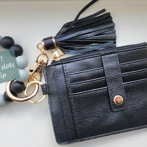 May include: A black cardholder with a gold zipper and snap closure, a black tassel keychain, and a beaded bracelet with the text "NEW! 9 card slots 1 zip". The cardholder has multiple card slots.