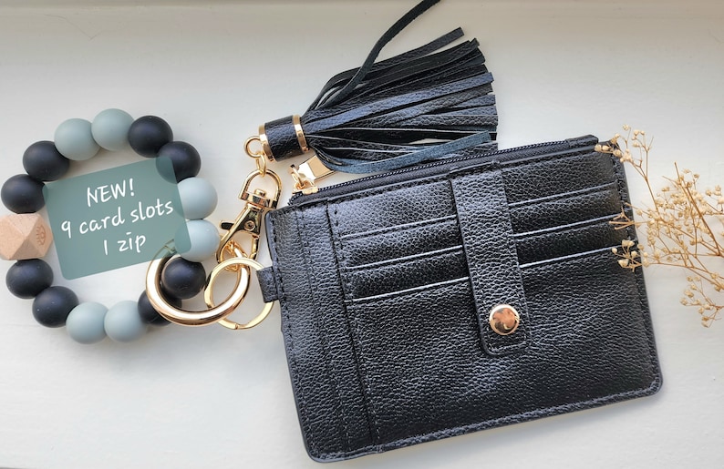 May include: A black cardholder with a gold zipper and snap closure, a black tassel keychain, and a beaded bracelet with the text "NEW! 9 card slots 1 zip". The cardholder has multiple card slots.