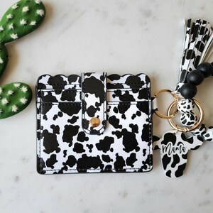 May include: A black and white cow print card holder with a gold snap closure. A matching cow print keychain with the word "Mama" is attached to a wristlet with black beads and a mint green silicone bracelet.