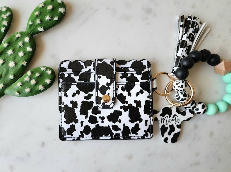 May include: A black and white cow print card holder with a gold snap closure. A matching cow print keychain with the word "Mama" is attached to a wristlet with black beads and a mint green silicone bracelet.