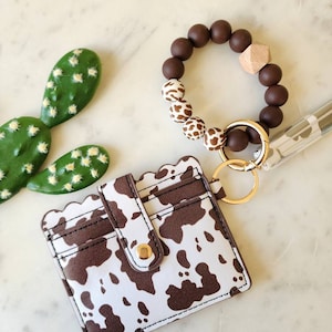 May include: A white and brown cow print card holder with a scalloped edge and a gold snap closure. A brown and white beaded bracelet with a wooden accent and gold hardware. A green cactus decoration is also present.