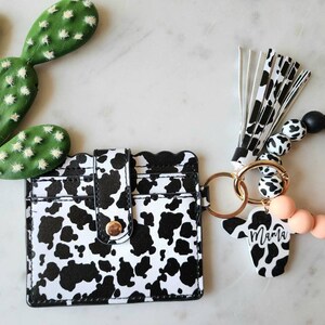 May include: A black and white cow print card holder with a gold snap closure and a matching keychain wristlet. The wristlet features black, white, and peach-colored beads, a wooden accent, and a cow-shaped charm with the word "Mama".