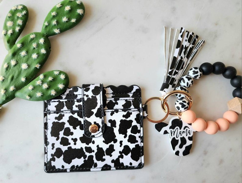May include: A black and white cow print card holder with a gold snap closure and a matching keychain wristlet. The wristlet features black, white, and peach-colored beads, a wooden accent, and a cow-shaped charm with the word "Mama".