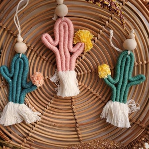 May include: Three handmade cactus ornaments with white tassels. The cacti are pink, green, and blue with yellow pom-poms on top. The ornaments are hanging from a woven wicker basket.