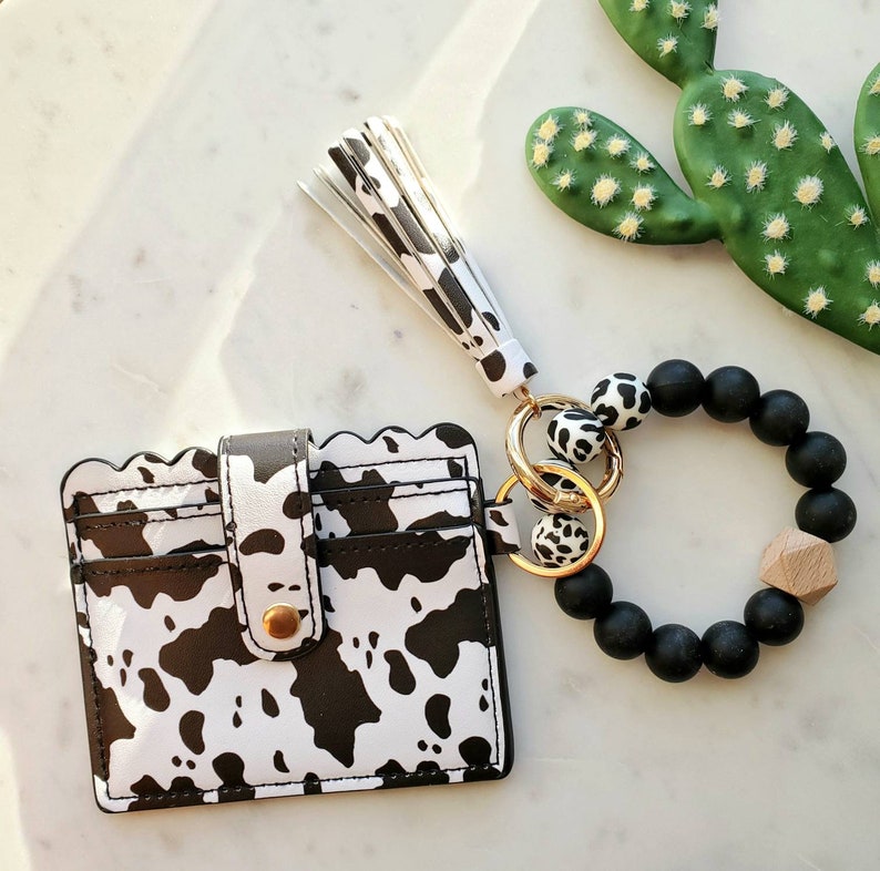 May include: A black and white cow print card holder with a scalloped edge, a matching tassel keychain, and a black beaded bracelet with a wooden accent. The card holder has a snap closure.