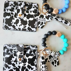 May include: Two cow-print cardholders with scalloped edges and snap closures are displayed. Two matching keychains with cow-print charms that read "Mama" and beaded bracelets in black, white, and blue or turquoise complete the set.