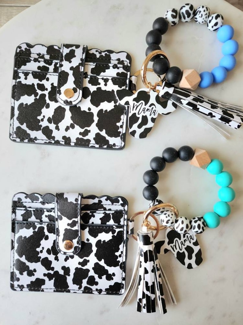 May include: Two cow-print cardholders with scalloped edges and snap closures are displayed. Two matching keychains with cow-print charms that read "Mama" and beaded bracelets in black, white, and blue or turquoise complete the set.