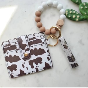 May include: A white and brown cow print cardholder with a scalloped edge, a matching lip balm holder, and a beaded keychain bracelet with white, brown, and wooden beads. The keychain has a gold clasp.