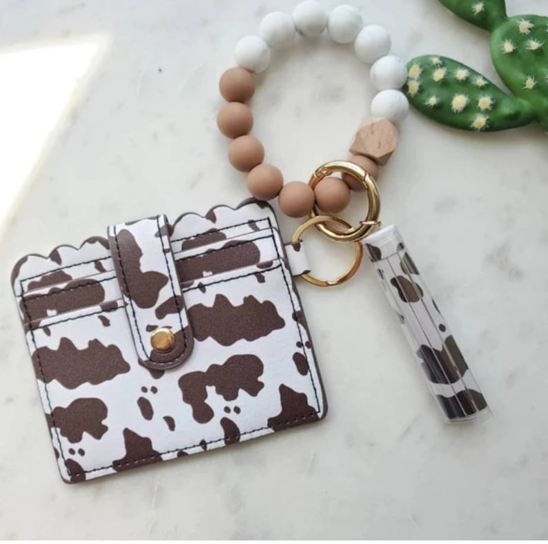 May include: A white and brown cow print cardholder with a scalloped edge, a matching lip balm holder, and a beaded keychain bracelet with white, brown, and wooden beads. The keychain has a gold clasp.
