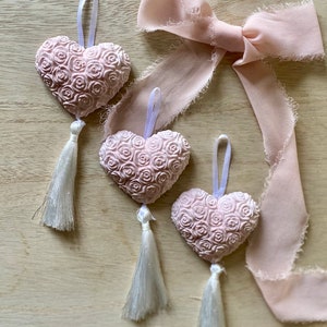 Op de afbeelding: Drie roze hartvormige ornamenten met een rozenpatroon, witte kwastjes en witte linten. Een lichtroze lint is in een strik geknoopt. De ornamenten liggen op een licht houten oppervlak.