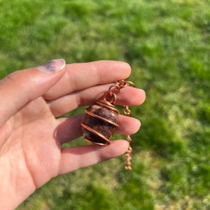 May include: A copper wire wrapped pendant featuring a brown and red stone. The pendant is attached to a copper chain.