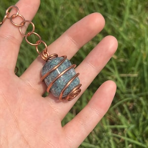 May include: A copper wire wrapped pendant featuring a green and blue speckled stone. The pendant is hanging from a copper chain.