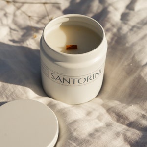 Peut inclure: Un pot de bougie blanc avec une mèche en bois et le mot "SANTORINI" imprimé sur le côté. Le pot est posé sur une surface en tissu blanc.