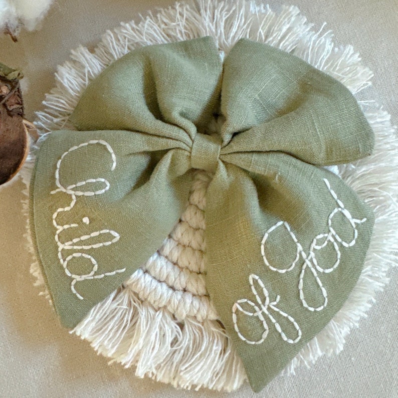 May include: A green fabric bow with white embroidery that reads "Child of God" on a white crocheted background.