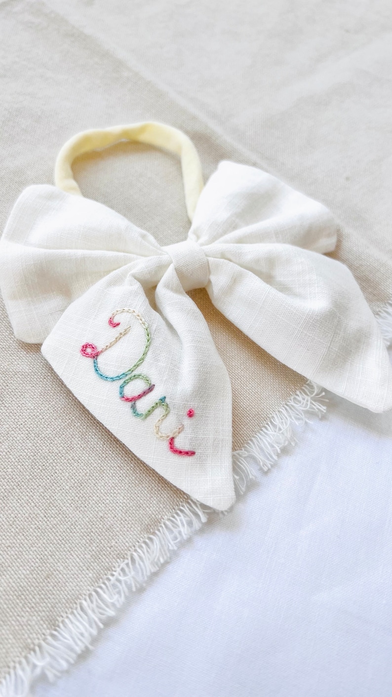May include: White fabric bow with a yellow elastic headband. The bow has the name "Dani" embroidered on it in colorful thread.