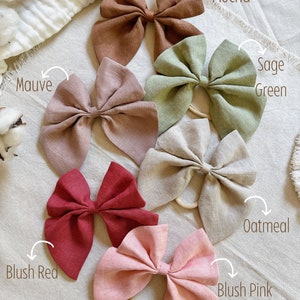 May include: Ten different colors of fabric hair bows, including blush pink, blush red, oatmeal, sage green, mauve, mocha. The bows are made of a soft, textured fabric and are tied with a knot in the center.